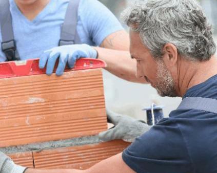 un artisan maçon cheveux gris et son jeune apprenti en train de monter un mur de parpaings