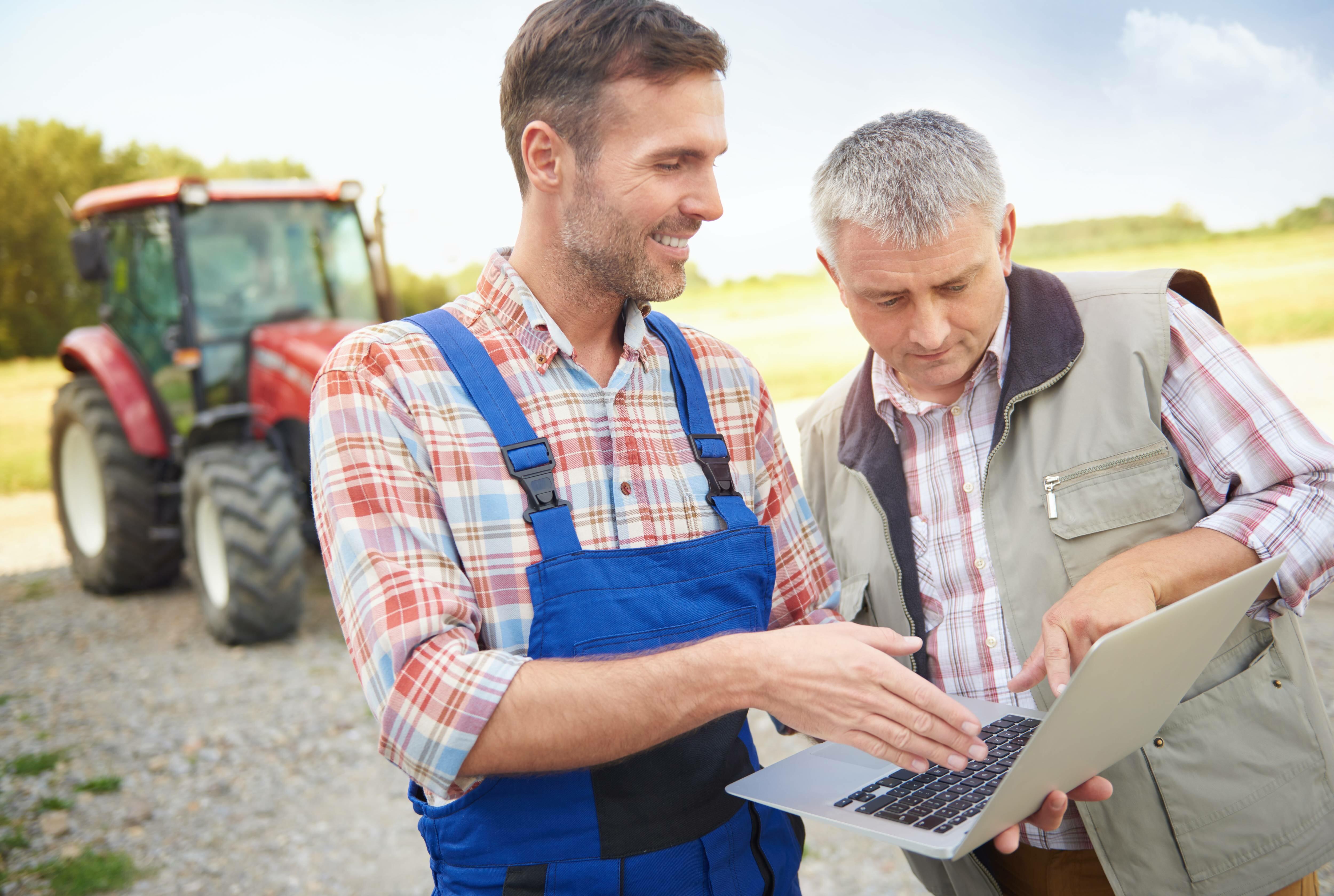 un jeune agriculteur en bleu de travail tenant un ordinateur erit un homme cheveux gris avec un tracteur en arrière-plan